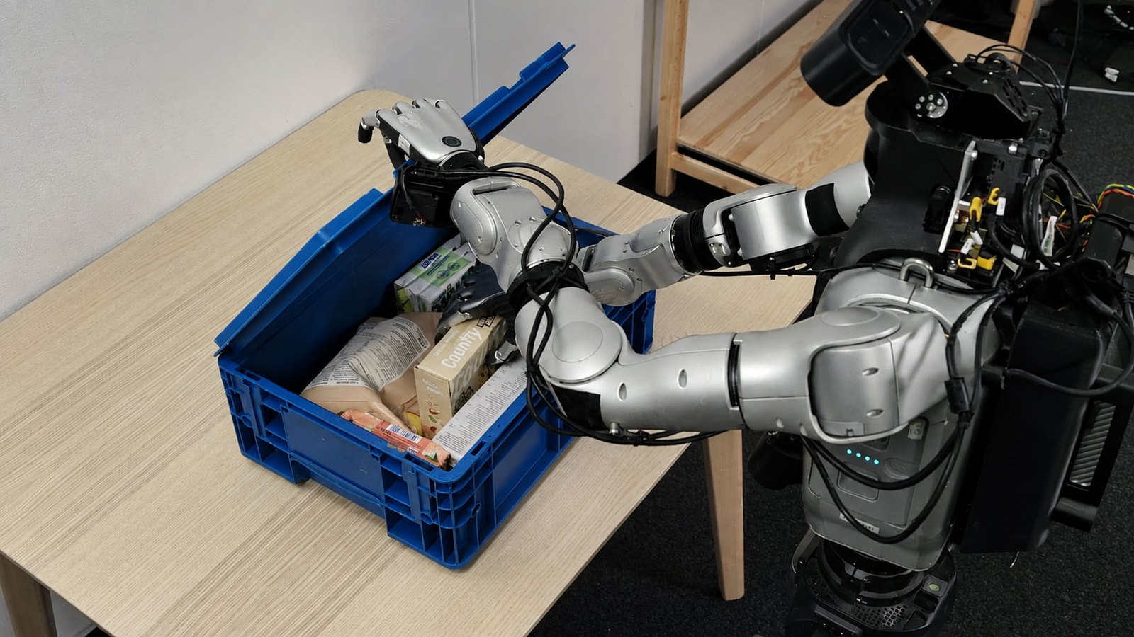A dual-arm robot reaching into a blue crate of groceries on a wooden table, one of Odyssey's robot manipulation demos